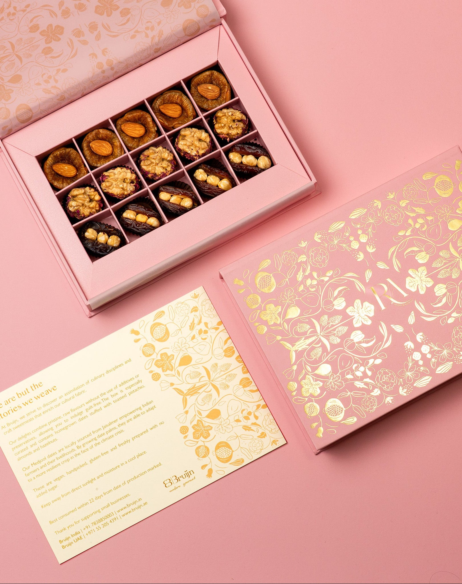 A fully customised box with bride and groom's logo in gold floral design, open to reveal chocolates, dried fruits like dates and figs on a pink background.