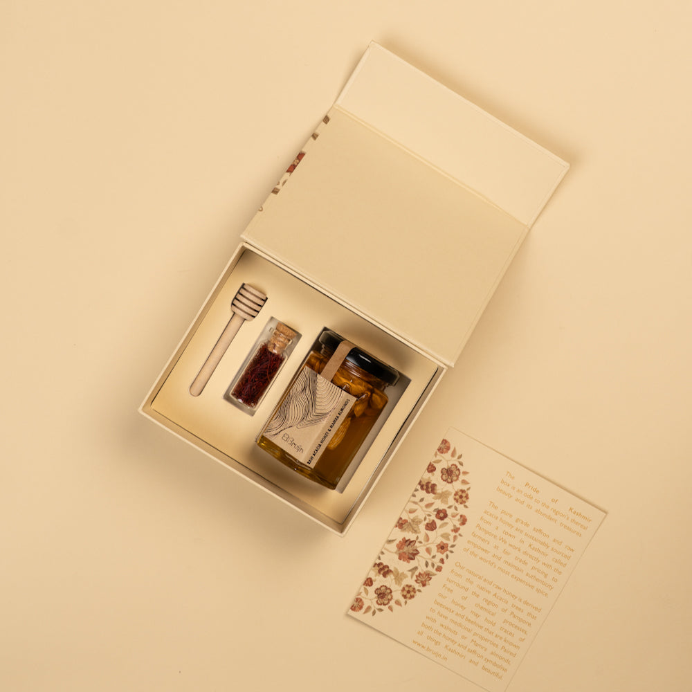 An open cream-colored gift box on a matching beige background, containing a small glass jar with amber liquid, a honey dipper, and a printed card with floral decoration and text.