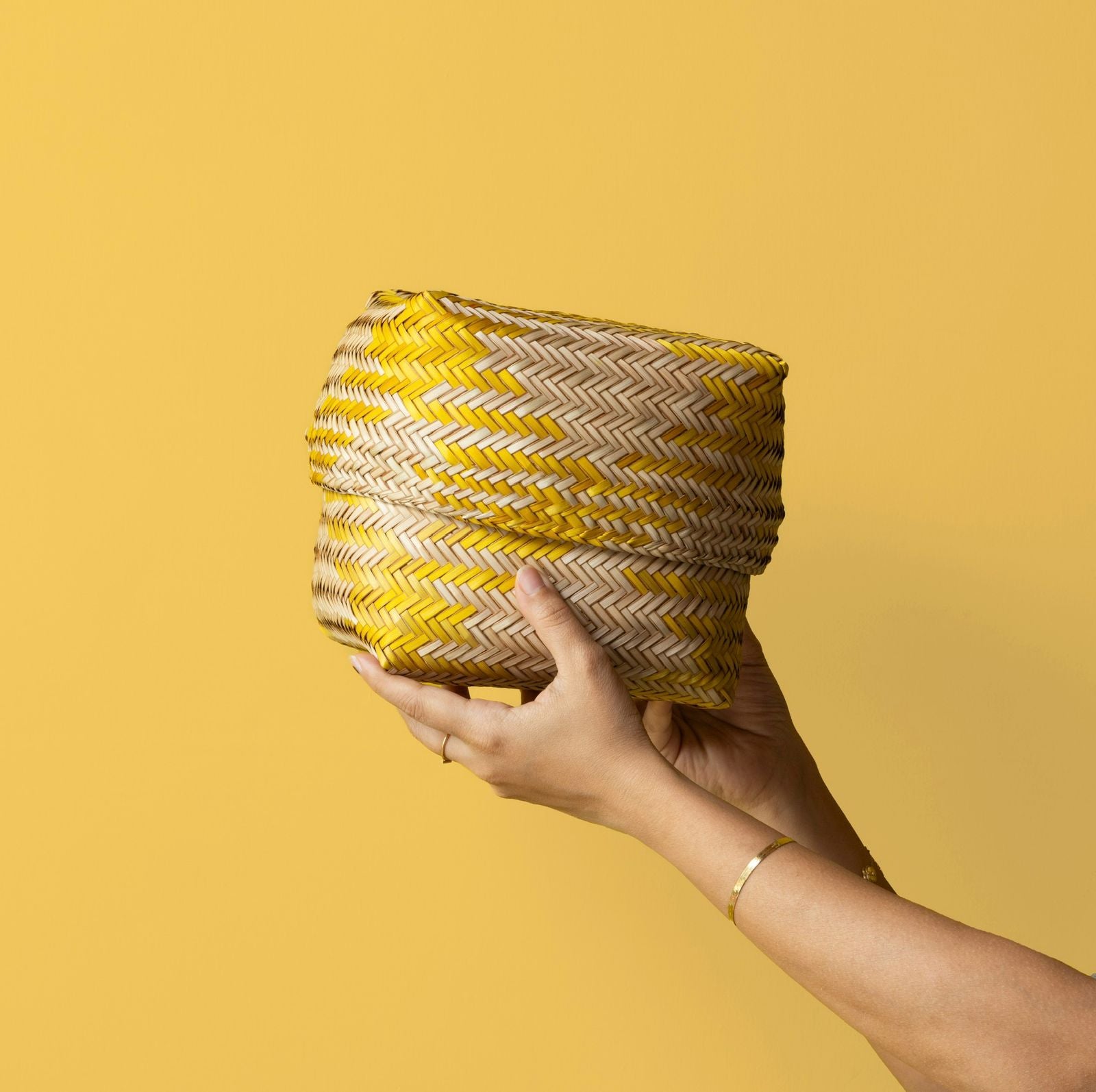 Hand holding a woven basket made from dried grass with yellow and beige pattern against a yellow background