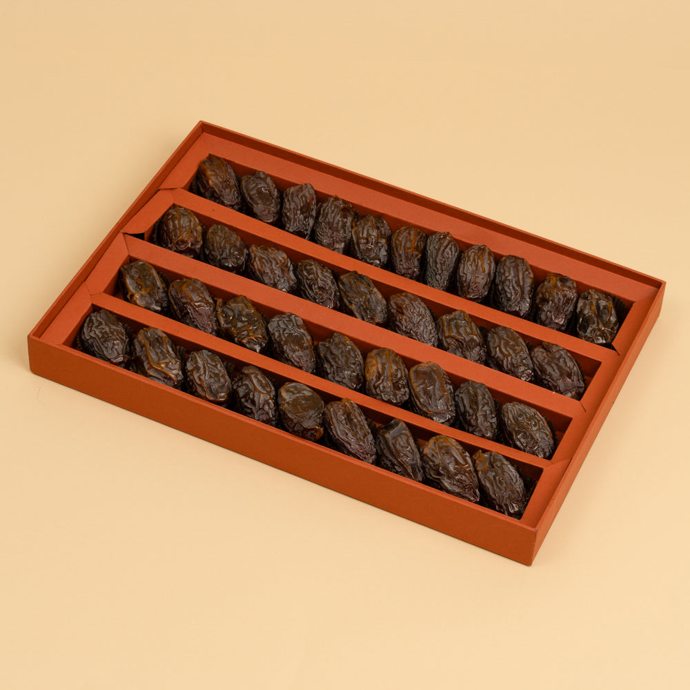 A rectangular reddish-brown gift box with five compartments, filled with dark brown dried dates, photographed on a solid beige background.