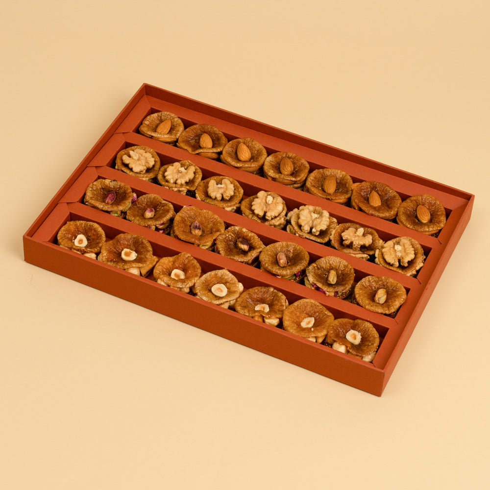 A rectangular reddish-brown gift box filled with assorted dried figs and nut snacks, arranged in rows on a warm beige background.