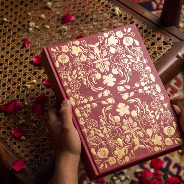 Red book with gold floral patterns held by a hand on a textured surface with flowers.