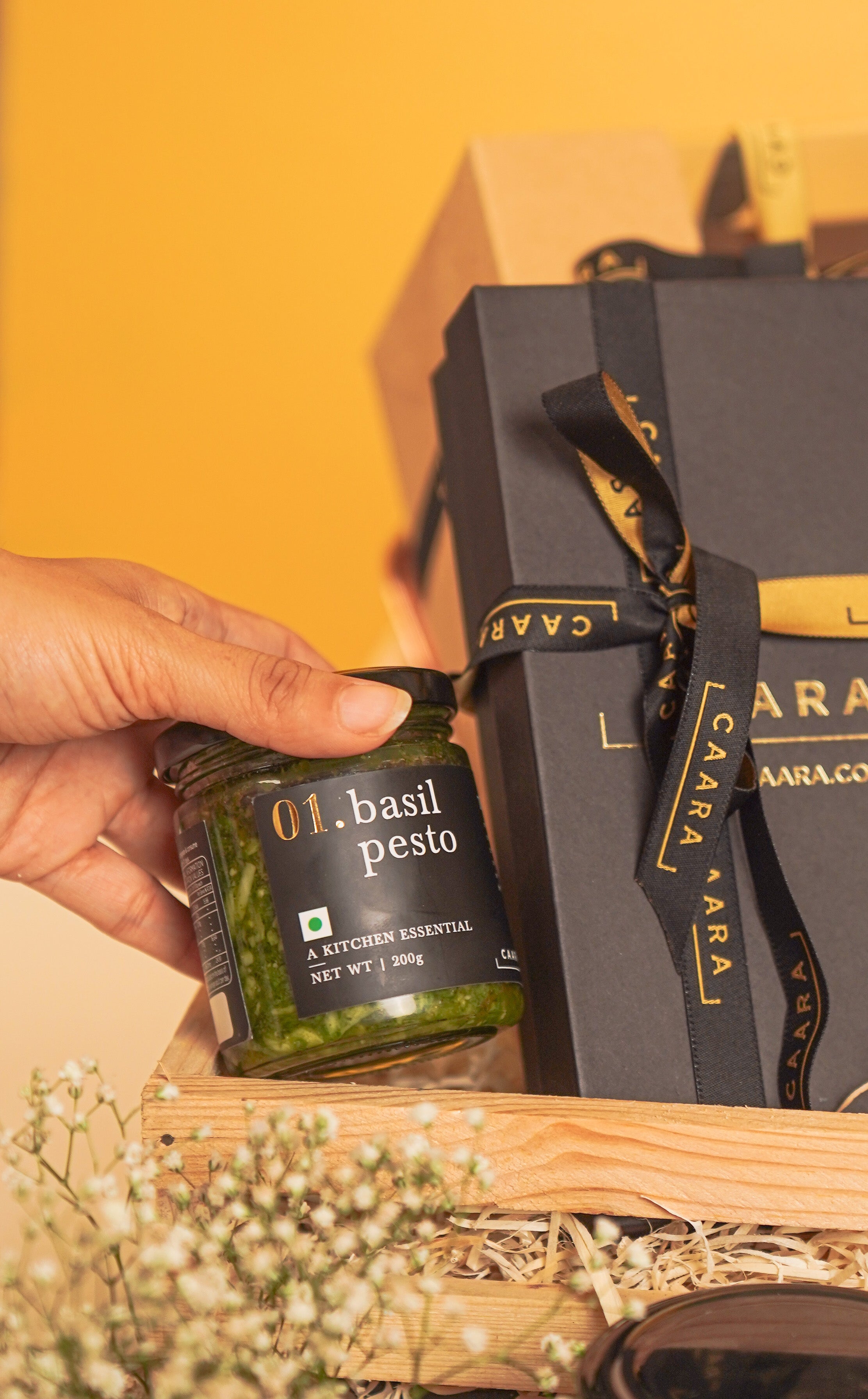 Hand holding a bottle of basil pesto next to a Caffarel gift box on a yellow background