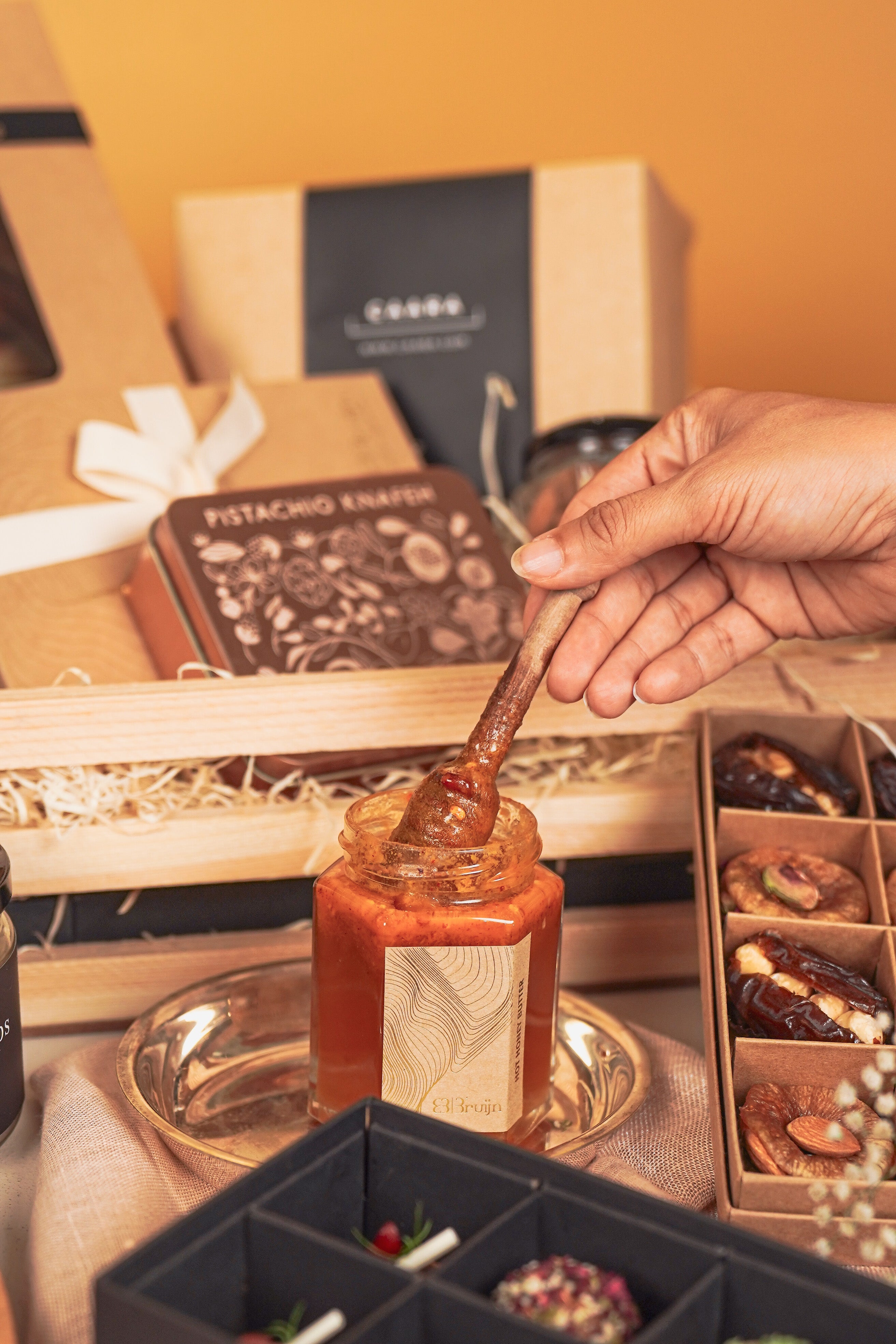 A honey dipper over a jar of honey with a gift box and snacks in the background.