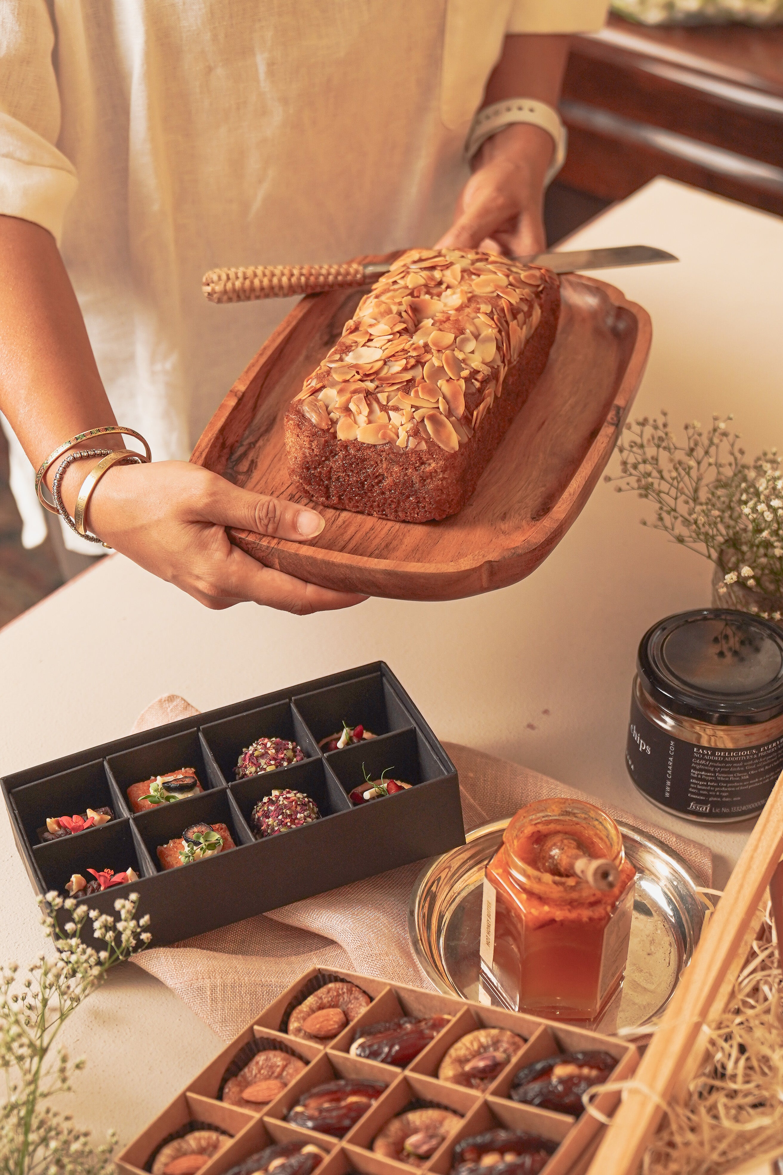 Person holding a loaf of bread on a wooden board with various food items and a candle in the background.