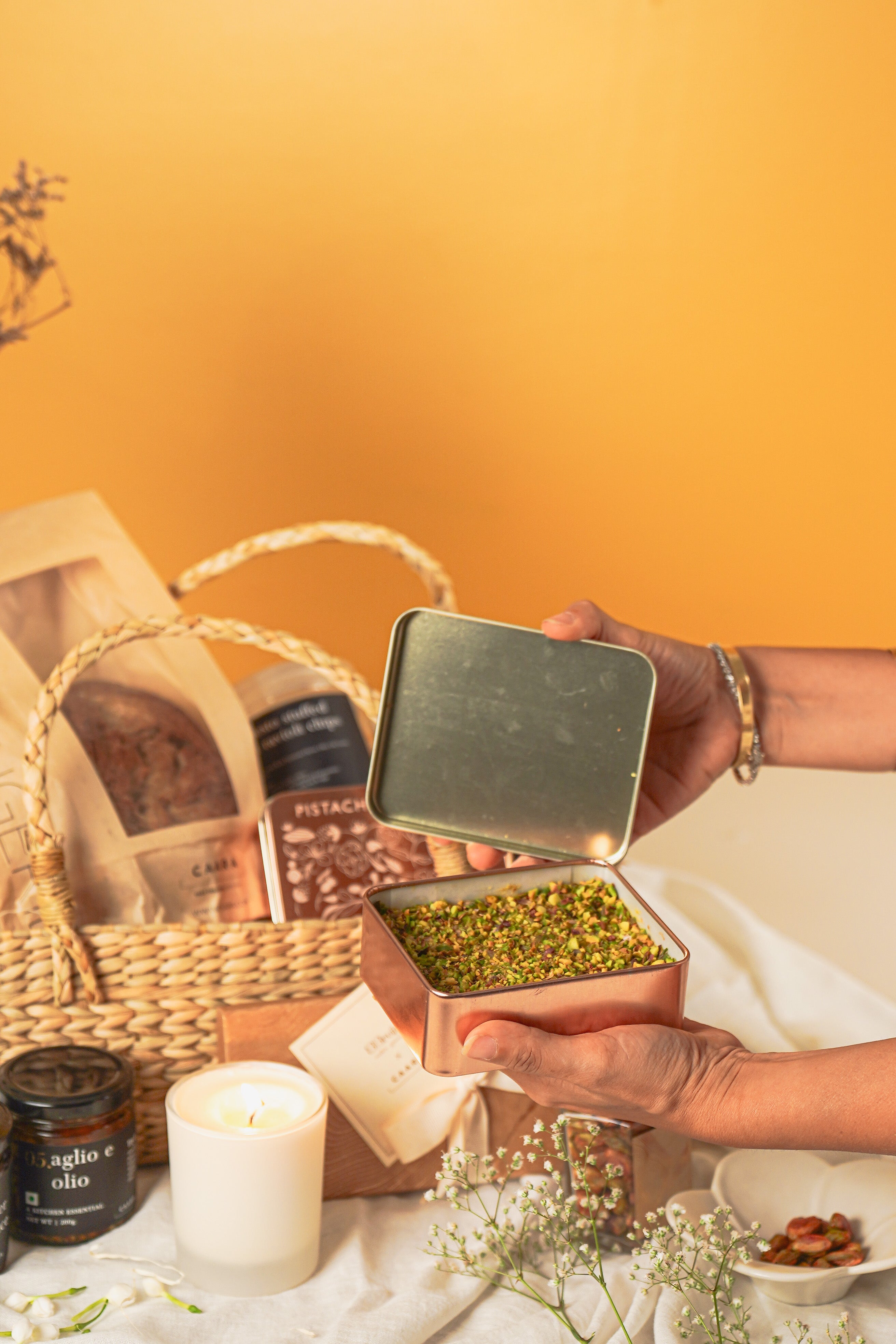  a small metal container filled with green herbs on a table with a basket and candles in the background.