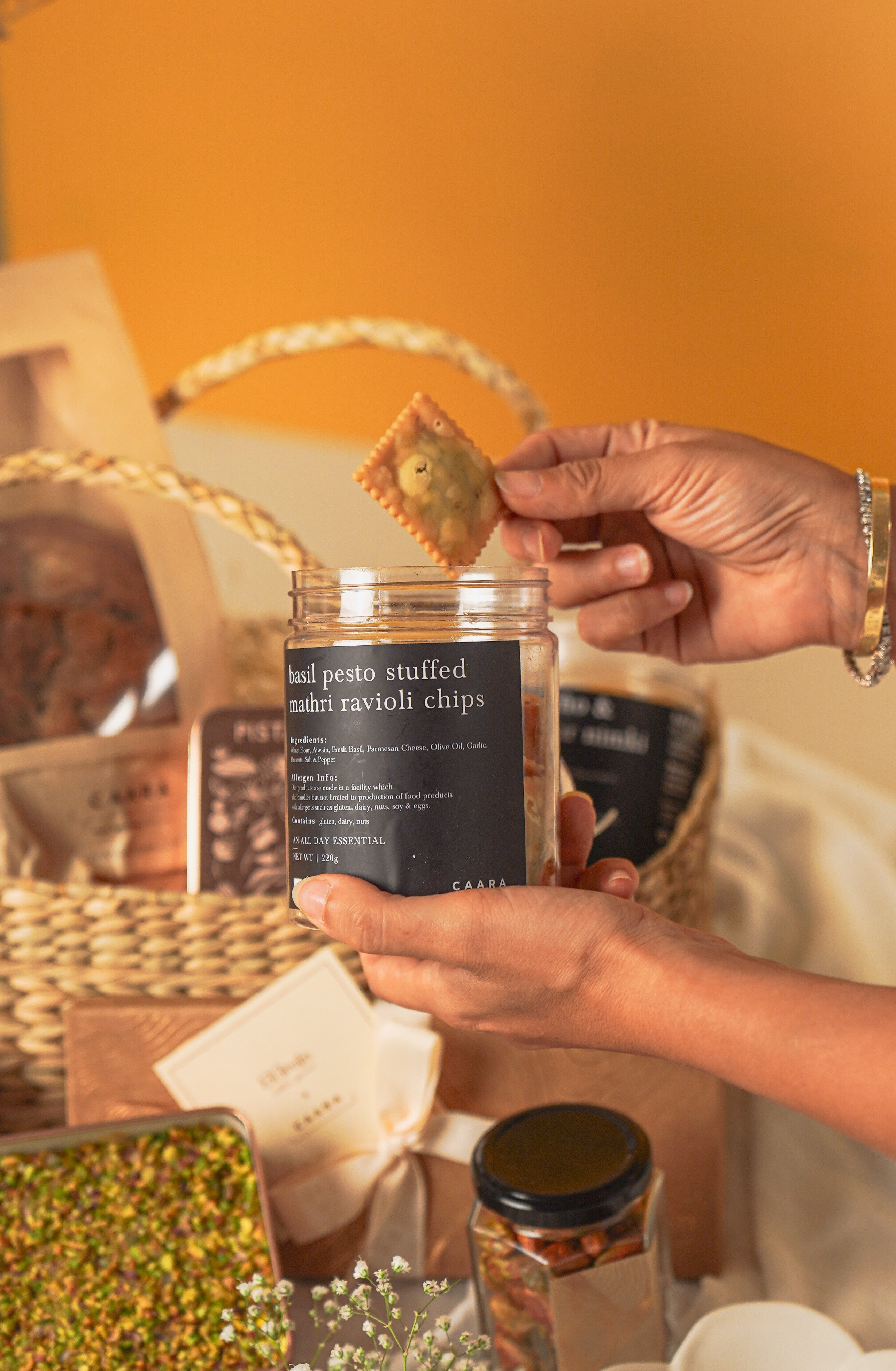 Person holding a jar of ravioli chips with a cracker, surrounded by gift items on a wooden surface.