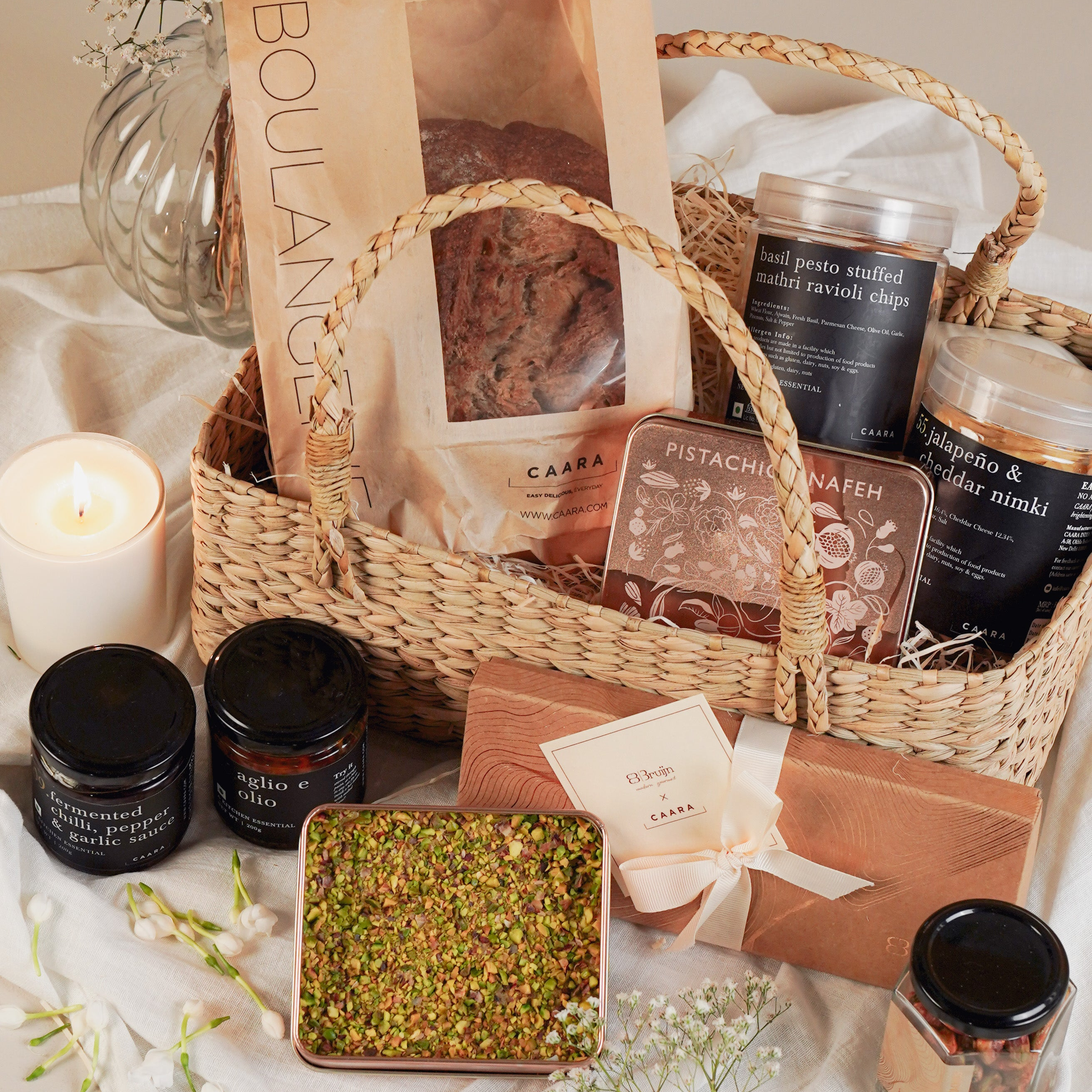 Wicker gift basket with various food items and a candle on a light surface.