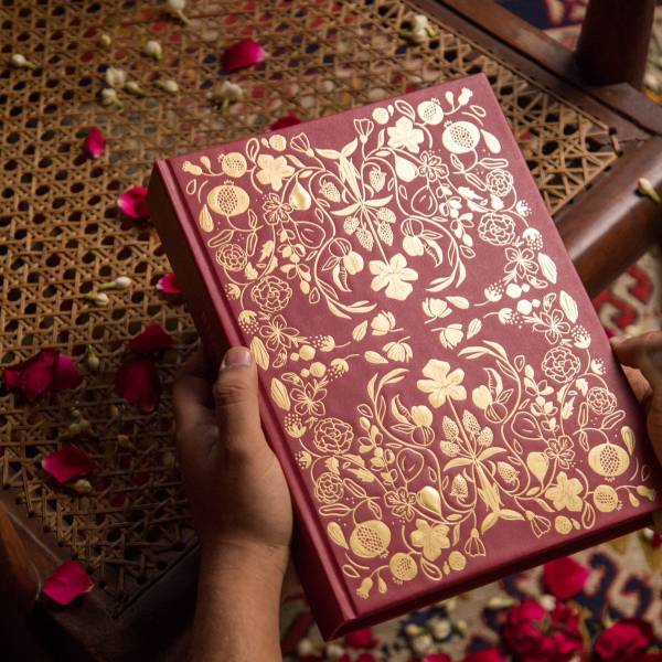 Red notebook with gold floral patterns held by a hand on a textured surface with rose petals.
